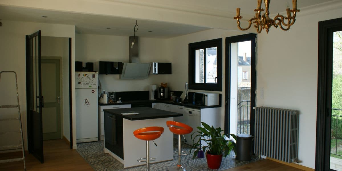 Cuisine moderne avec îlot central, tabourets orange, placards blancs et fenêtres à cadres noirs.