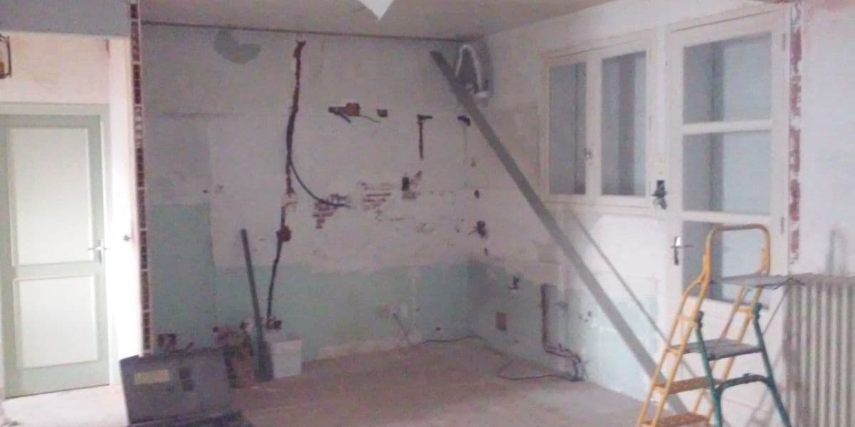Chantier de rénovation intérieure avec murs dénudés, fils électriques apparents, escabeau et outils au sol.