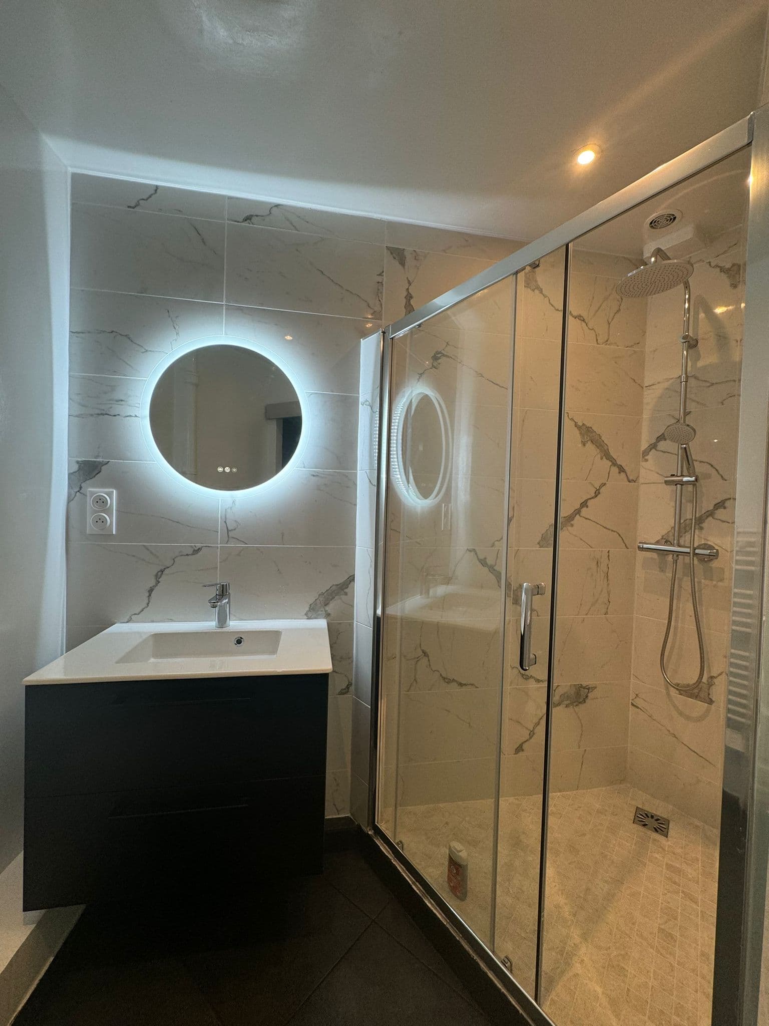 Salle de bain moderne avec meuble noir, miroir rond rétroéclairé et douche vitrée en marbre.